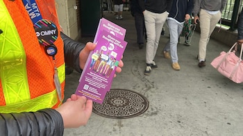 Uma pessoa com um colete de segurança laranja segura um livreto roxo da pesquisa MTA Customers Count; pessoas andando ao fundo