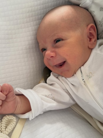 Primer plano de un bebé sonriente, acostado de lado en una cuna blanca. El bebé, con mameluco blanco, mira hacia la derecha y tiene la boca abierta en una amplia sonrisa