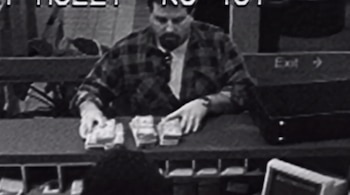 Un hombre barbudo con camisa a cuadros, tres montones de billetes sobre un mostrador, una persona sentada, una señal de salida, un bolso negro y un monitor.