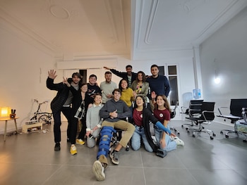 Un grupo de doce personas, once de pie y una sentada con férula en la pierna, sonríen en una habitación blanca con techo decorado y suelo gris