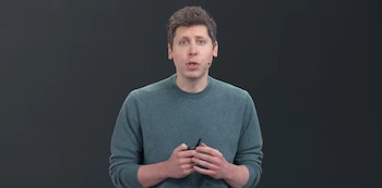 Sam Altman viste un suéter verde frente a un fondo gris y sostiene un pequeño dispositivo negro entre las manos