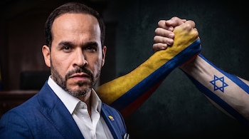Un hombre de traje oscuro y barba mira al frente. Dos brazos con las banderas de Colombia e Israel realizan un apretón de manos.