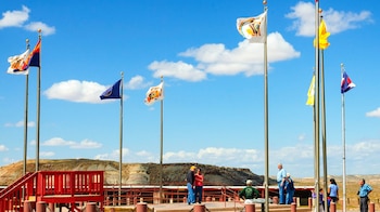 Varias banderas ondean en postes altos sobre un mirador de madera roja con personas y un paisaje desértico de colinas bajo un cielo azul nublado