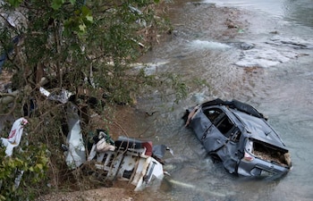Un coche gris muy dañado está parcialmente sumergido en un río o arroyo. A la izquierda, un árbol y otros escombros en la orilla