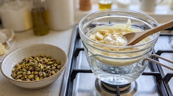 Un recipiente con pistachos pelados al lado de un bol de vidrio con chocolate blanco derretido sobre una hornalla de cocina.