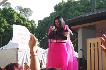 Performance M, con un vestido rosa brillante y guantes negros, sonríe en un escenario al aire libre. Sostiene un micrófono y un teléfono