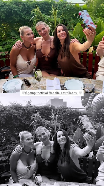 Tres mujeres sentadas a una mesa de madera con platos y copas en un restaurante al aire libre, con plantas verdes de fondo. Una mujer toma una selfie con un teléfono