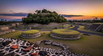 Vista panorámica del sitio arqueológico de Cuicuilco al atardecer, con la pirámide circular, estructuras de piedra, césped y senderos. Edificios modernos se ven a lo lejos bajo un cielo vibrante.