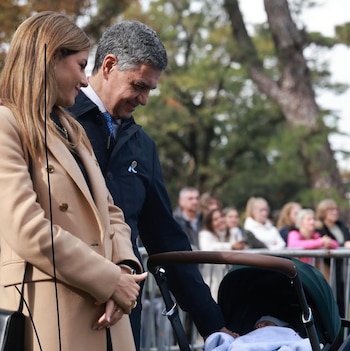 Una mujer con abrigo camel y un hombre de chaqueta azul observan a un bebé en un cochecito oscuro en un evento al aire libre con espectadores de fondo