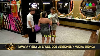 Tres personas en un baño. Una mujer de vestido brillante plateado y otra de vestido negro strapless discuten. Un hombre con camiseta blanca observa