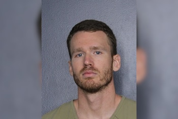 Fotografía policial de un hombre joven con barba corta y camiseta verde clara frente a una pared gris