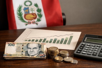 Un billete de cien soles, monedas, una calculadora digital y un gráfico en papel sobre una mesa de madera oscura. La bandera de Perú está desenfocada al fondo.