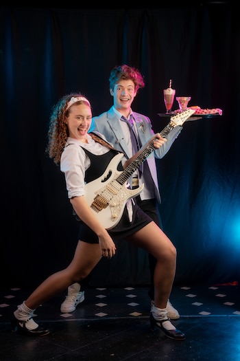 Una mujer sostiene una guitarra eléctrica blanca mientras un hombre detrás lleva una bandeja con una bebida y aperitivos