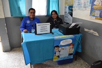 Un hombre y una mujer sentados detrás de una mesa azul con una computadora portátil, una impresora y afiches informativos