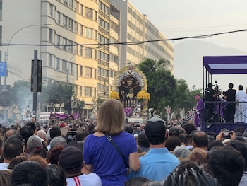 Multitud reunida en una avenida frente al anda con la imagen del Señor de los Milagros y un estrado con un sacerdote vestido de morado