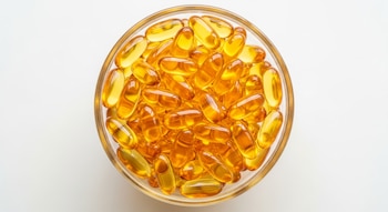 Vista superior de un cuenco transparente lleno de cápsulas blandas de color amarillo dorado, brillantes y translúcidas, sobre un fondo blanco iluminado.