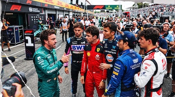 Pilotos con monos de carreras de automovilismo conversan de pie en el área de garajes ante fotógrafos y miembros del equipo.