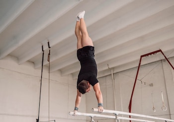 Un hombre con ropa deportiva negra realiza una vertical apoyando las manos en dos barras paralelas dentro de un gimnasio