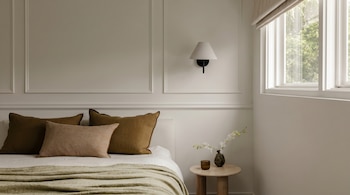 Una habitación de estilo minimalista con una cama vestida con almohadas marrones y una manta verde oliva, una mesita de noche de madera y una ventana grande.