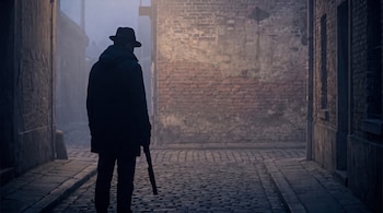 Silueta posterior de un hombre con sombrero y abrigo que sostiene un arma en un callejón empedrado con muros de ladrillo y niebla.