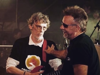 Luis Alberto Spinetta sostiene una guitarra mientras León Gieco le apoya una mano en el hombro en un escenario iluminado