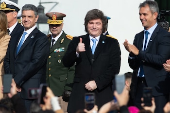 Presidente Javier Milei con cabello castaño y abrigo negro, sonriendo y haciendo un gesto de aprobación con el pulgar. A su lado, hombres en traje y uniforme militar. Fondo claro, manos de personas con teléfonos