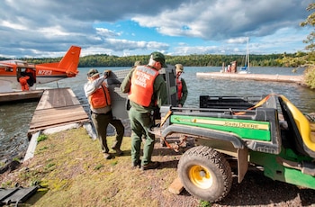 Dos guardaparques con chalecos salvavidas naranjas cargan una jaula metálica desde un muelle hacia un vehículo John Deere Gator. Un hidroavión naranja está a la izquierda