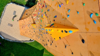 Vista aérea de la Torre Excalibur, una pared de escalada al aire libre de color naranja con cientos de agarres multicolores, junto a una zona de grava y césped verde.