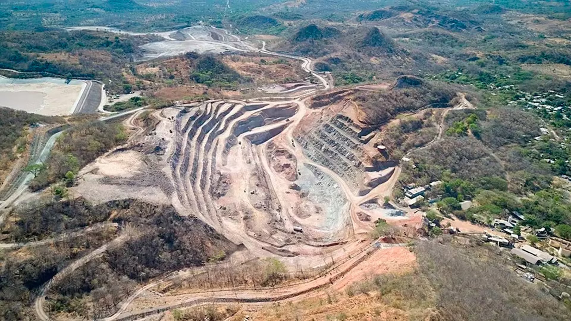 Vista aérea de una gran mina de oro a cielo abierto con sus terrazas excavadas, carreteras internas y una presa de relaves adyacente, mostrando el impacto en el paisaje.