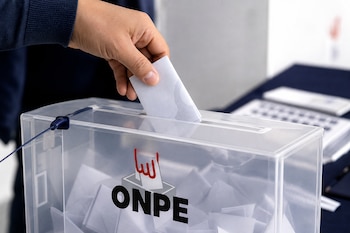 Una mano deposita una cédula de papel en una urna transparente con el logo de la ONPE llena de votos doblados.