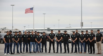 Un grupo de diecisiete hombres con camisetas negras y gorras, algunos con gafas de sol, posan en fila en un aparcamiento. Al fondo, vehículos, postes de luz y la bandera de Estados Unidos.