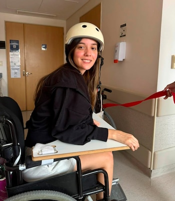 Una mujer joven, Manuela Lori, con cabello castaño largo y un casco color crema, sonríe a la cámara mientras está sentada en una silla de ruedas en un hospital