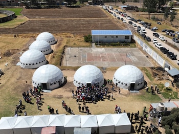 Vista aérea de una explanada con seis domos blancos, varias carpas, personas, vehículos, una cancha deportiva y campos de cultivo