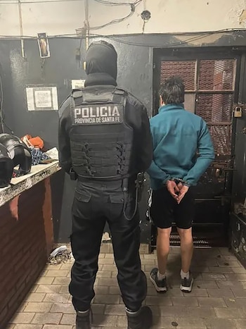 Vista trasera de un oficial de policía de Santa Fe con chaleco negro frente a un hombre esposado de espaldas, con chaqueta turquesa, en un recinto interior