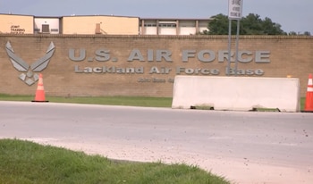Letrero de la Base Aérea Lackland, logo de la Fuerza Aérea de Estados Unidos, muro de ladrillo, dos conos, barrera de hormigón, carretera y pasto