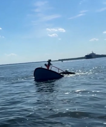 Una lancha de rescate parcialmente hundida con una persona a bordo, visible desde el agua. Se observa un barco lejano y cielo azul