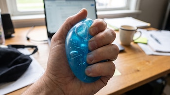Una mano aprieta una pelota antiestrés azul transparente. Se ve un portátil, documentos, una taza y una ventana en el fondo.