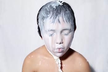Un niño con el torso descubierto y los ojos cerrados tiene un líquido blanco espeso vertido sobre la cabeza y la cara frente a un fondo blanco