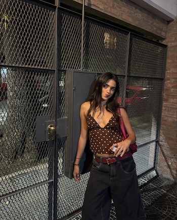 Maia Reficco, una joven de cabello oscuro y liso, posa con una blusa de lunares y pantalones negros anchos frente a una reja metálica en la noche