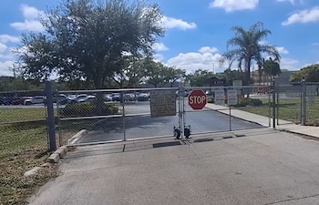 Reja de malla metálica cerrada con cartel de 'STOP' y aviso de seguridad. Al fondo, un estacionamiento con autos y árboles bajo cielo azul