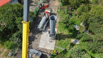Vista aérea de una grúa amarilla y operarios en el montaje de un poste cilíndrico metálico junto a un camión y áreas verdes