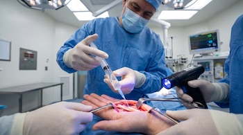 Cirujano en quirófano aplicando biopolímero con jeringa sobre nervio seccionado en mano de paciente, asistido por luz azul y otros instrumentos quirúrgicos.