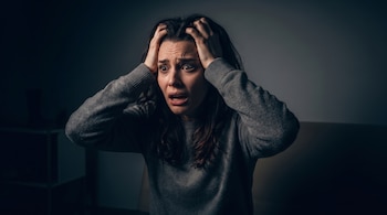 Una mujer con cabello oscuro y jersey gris se sujeta la cabeza con ambas manos. Su boca está ligeramente abierta y sus ojos miran al frente.