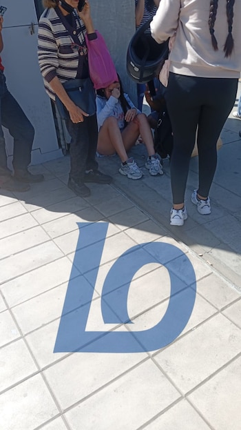 Joven sentada en acera con rodilla herida y bolsa rosa en la cabeza, asistida por adultos. Gran 'LO' azul pintado en el suelo de baldosas