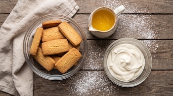 Un paño de tela. Un bol con galletas rectangulares. Una jarra con manteca derretida. Un bol con crema de leche. Azúcar impalpable y una mesa de madera.