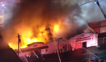 Un edificio nocturno envuelto en grandes llamas y humo denso recibe chorros de agua desde mangueras de bomberos