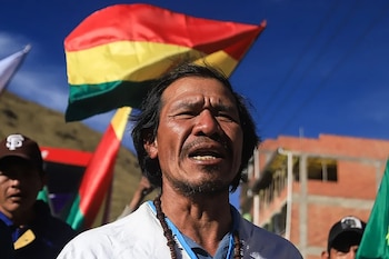 Primer plano de un hombre indígena de mediana edad con cabello oscuro, barba, collar de cuentas y camiseta blanca, con una bandera de Bolivia al fondo