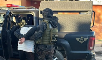 Un agente táctico sujeta a un hombre esposado por la espalda junto a un vehículo policial