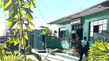 Vista exterior de una comisaría verde con un cartel que dice "Policía Nacional". Un oficial de policía camina hacia la entrada. Hay plantas y árboles en primer plano