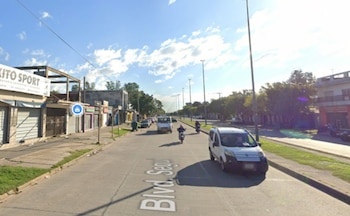 Calle concurrida con edificios de tiendas a la izquierda, vehículos como una camioneta blanca y motos en movimiento, y árboles a la derecha, bajo un cielo azul